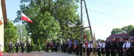Zdjęcie przedstawia ustawione w rzędzie poczty flagowe OSP oraz druhów OSP stojących przed remizą