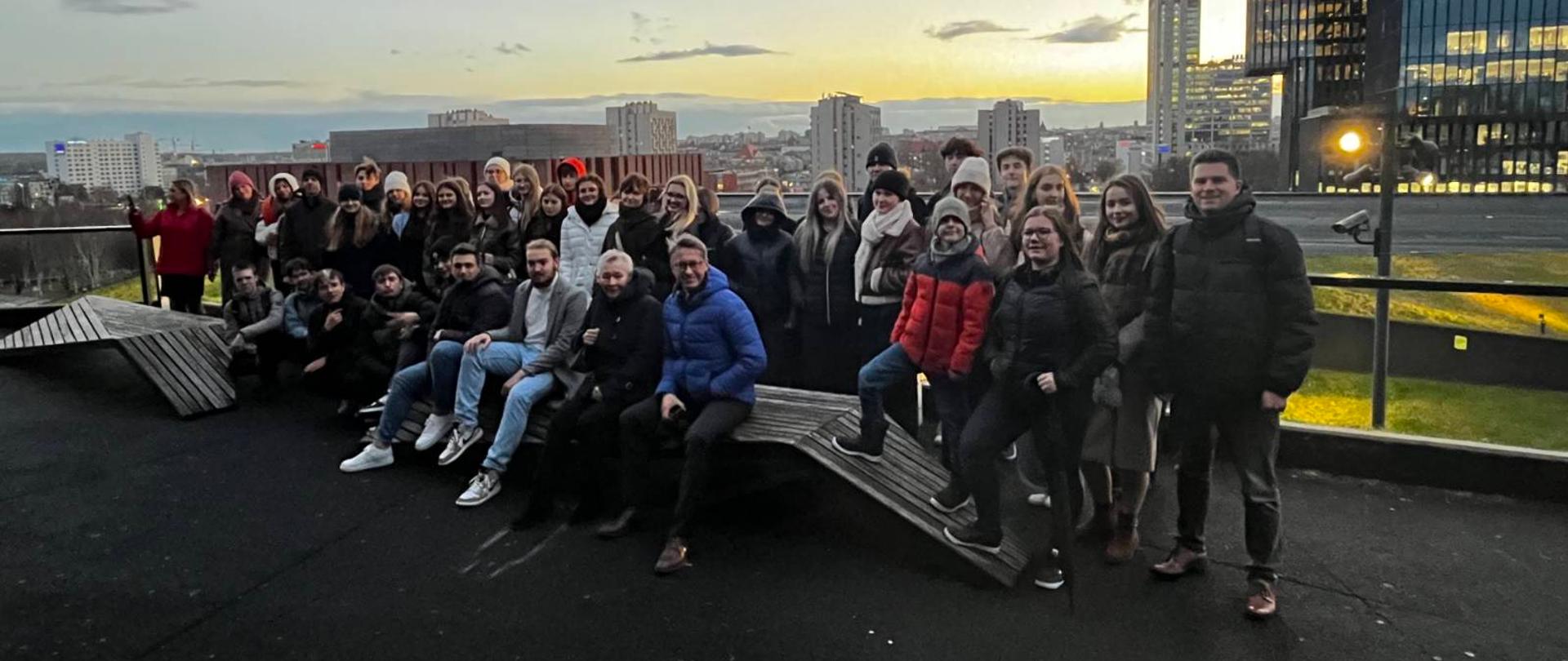 Grupa ludzi pozująca do zdjęcia na tarasie widokowym z panoramą miasta w tle. Widać nowoczesne budynki, wieżowce oraz rozświetlone niebo o zmierzchu. Osoby ubrane są w zimowe kurtki i czapki, co wskazuje na chłodną porę roku.