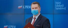 Dzień Edukacji Narodowej, minister Przemysław Czarnek trzyma teczkę. w tle niebieska plansza z napisem KPRM i DZień Edukacji Narodowej 