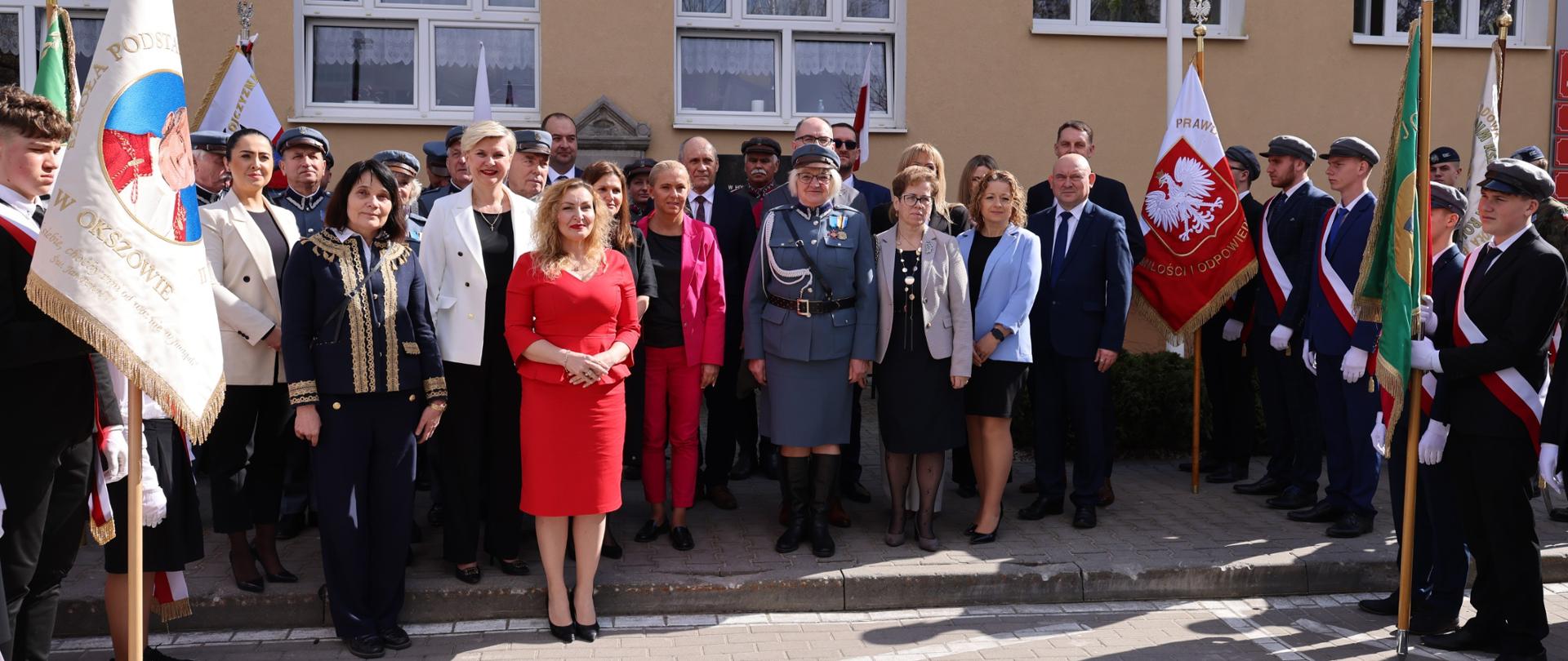 Osoby biorące udział w uroczystości stoją przed budynkiem, ustawione w kilku rzędach. Po obu stronach widoczne są poczty sztandarowe z flagami i sztandarami w różnych barwach. Na środku stoją osoby w strojach formalnych i mundurowych, a cała scena oświetlona jest światłem dziennym.
