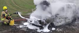 na zdjęciu spalony całkowicie samochód z otwartą maska silnika obok niego strażak podaje prąd piany z żółtego pojemnika na palący się pojazd widać kłęby białego dymu w oddali pole uprawne