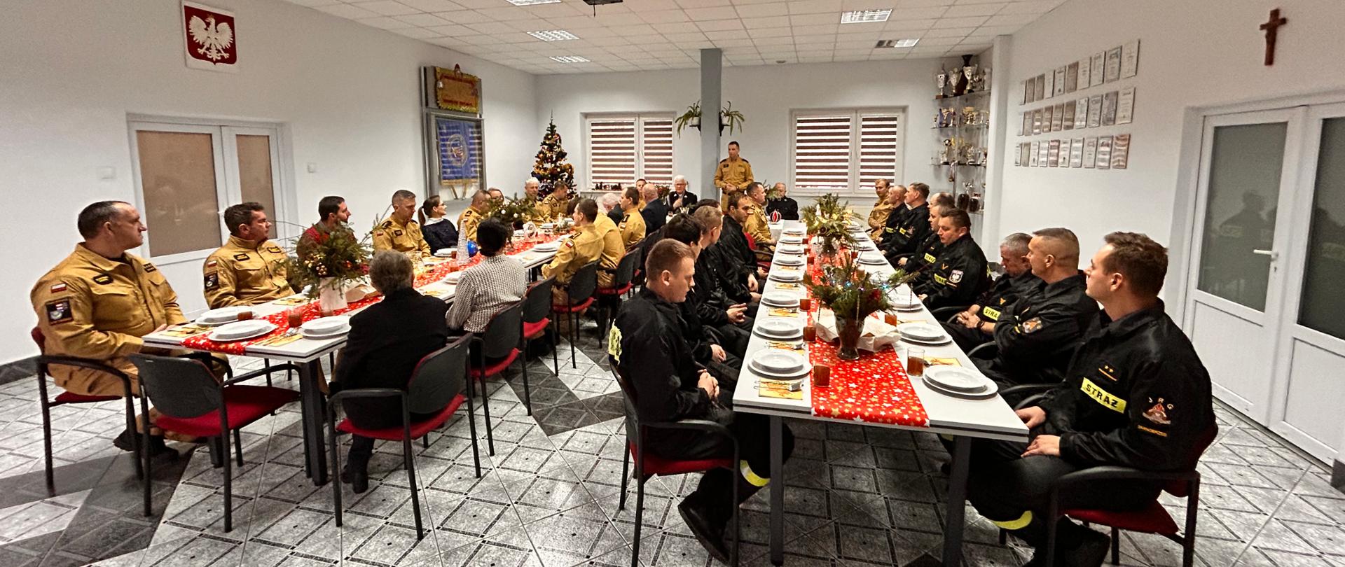 w pomieszczeniu strażacy siedzący przy udekorowanych świątecznie stołach. Jeden ze strażaków stoi i przemawia do pozostałych. W pomieszczeniu widoczna udekorowana choinka. 