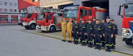 Strażacy na zbiórce oddają honory dla zmarłego strażaka na tle samochodów przed jednostką. 