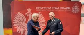 Na zdjęciu widzimy dwóch mężczyzn podających sobie ręce. Mężczyzna z prawej strony kadru to strażak, ubrany w mundur galowy kolor czarnego. Mężczyzna z lewej to cywil ubrany w granatową bluzę oraz oraz spodnie. Mężczyźni w lewych dłoniach trzymają dokumenty. W tle widzimy czerwony baner koloru czerwonego z godłem polski oraz napisem Państwowa Straż Pożarna. 