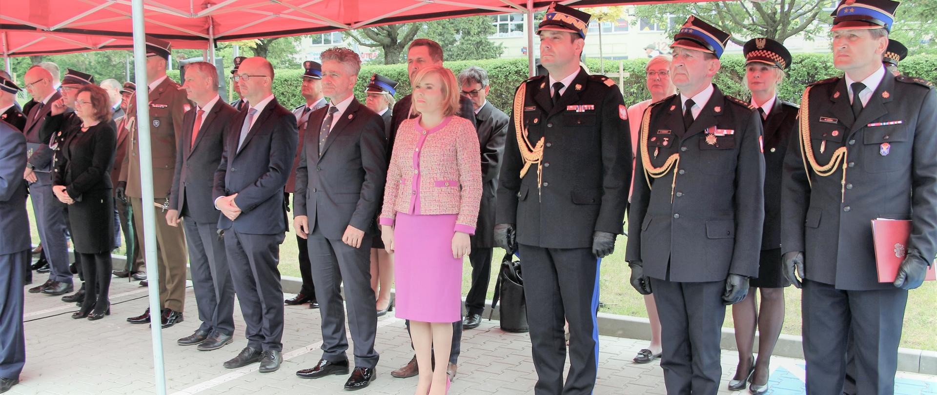 Pod czerwonym namiotem stoją zaproszeni goście wraz z gospodarzami uroczystości.