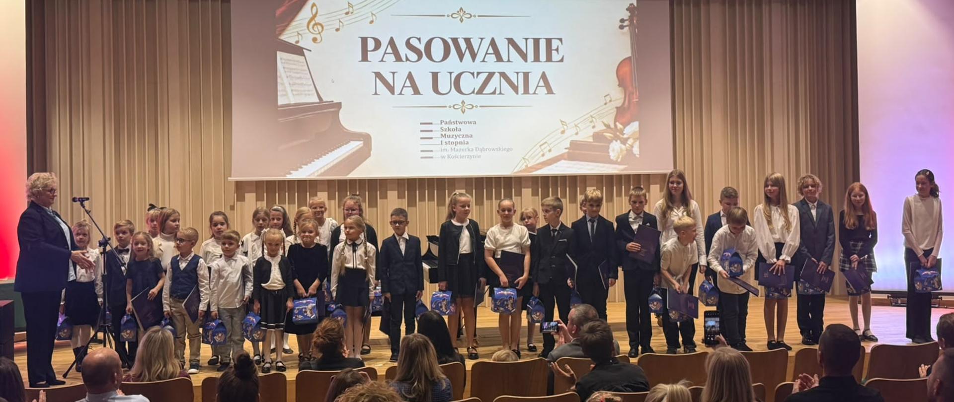 Na scenie duża grupa odświętnie ubranych dzieci, którzy stoją w wokół czarnego fortepianu, na którym gra mężczyzna w czarnej marynarce. Tło obrazka w odcieniach beżu. Zdjęcie zostało wykonane podczas koncertu z okazji Dnia Edukacji Narodowej połączonego z uroczystym ślubowaniem uczniów klas pierwszych. 