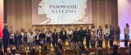 Na scenie duża grupa odświętnie ubranych dzieci, którzy stoją w wokół czarnego fortepianu, na którym gra mężczyzna w czarnej marynarce. Tło obrazka w odcieniach beżu. Zdjęcie zostało wykonane podczas koncertu z okazji Dnia Edukacji Narodowej połączonego z uroczystym ślubowaniem uczniów klas pierwszych. 
