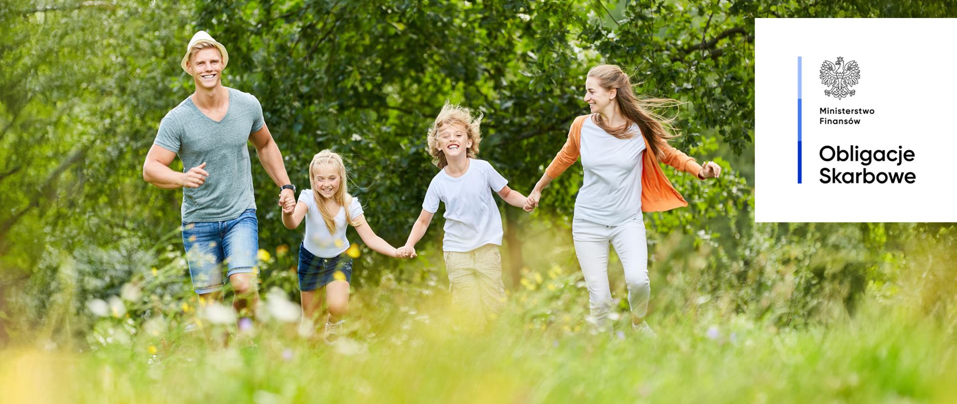 Cztery osoby biegną trzymając się za ręce, w otoczeniu zielonych drzew, w słoneczny dzień. Ministerstwo Finansów, Obligacje Skarbowe.