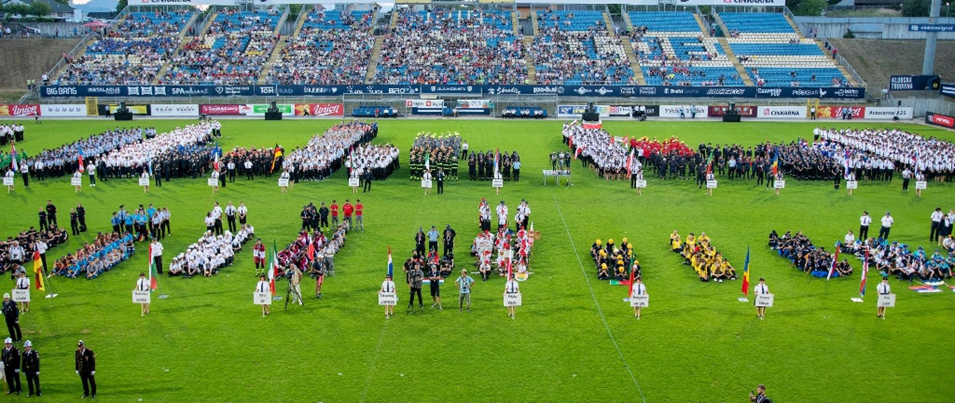 na stadione, na zielonej trawie stoją grupy ludzi, widok z daleka, za nimi trybuna z widzami, zawody 