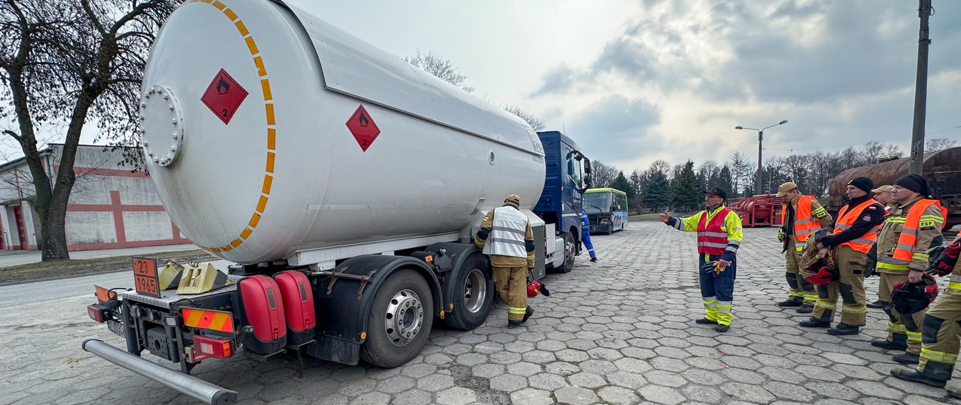 Widok z prawego boku na ciągnik siodłowy z podpiętą cysterną, po prawej stojący instruktor wraz z grupą uczestników warsztatów