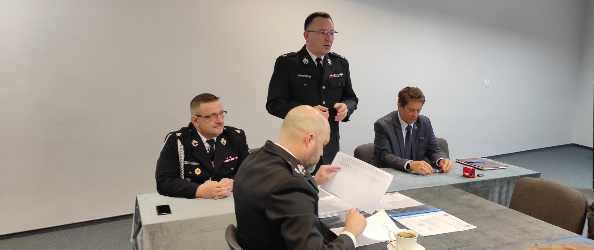 Zebranie, spotkanie na sali, białe ściany. Za biurkiem trzech strażaków w mundurach wyjściowych oraz starosta braniewski w garniturze.