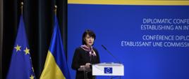 Na zdjęciu jest kobieta, która ma na sobie ciemne ubrania. Stoi przy mównicy. Mówi do dwóch mikrofonów. Za nią są dwie flagi - Unii Europejskiej i Ukrainy.