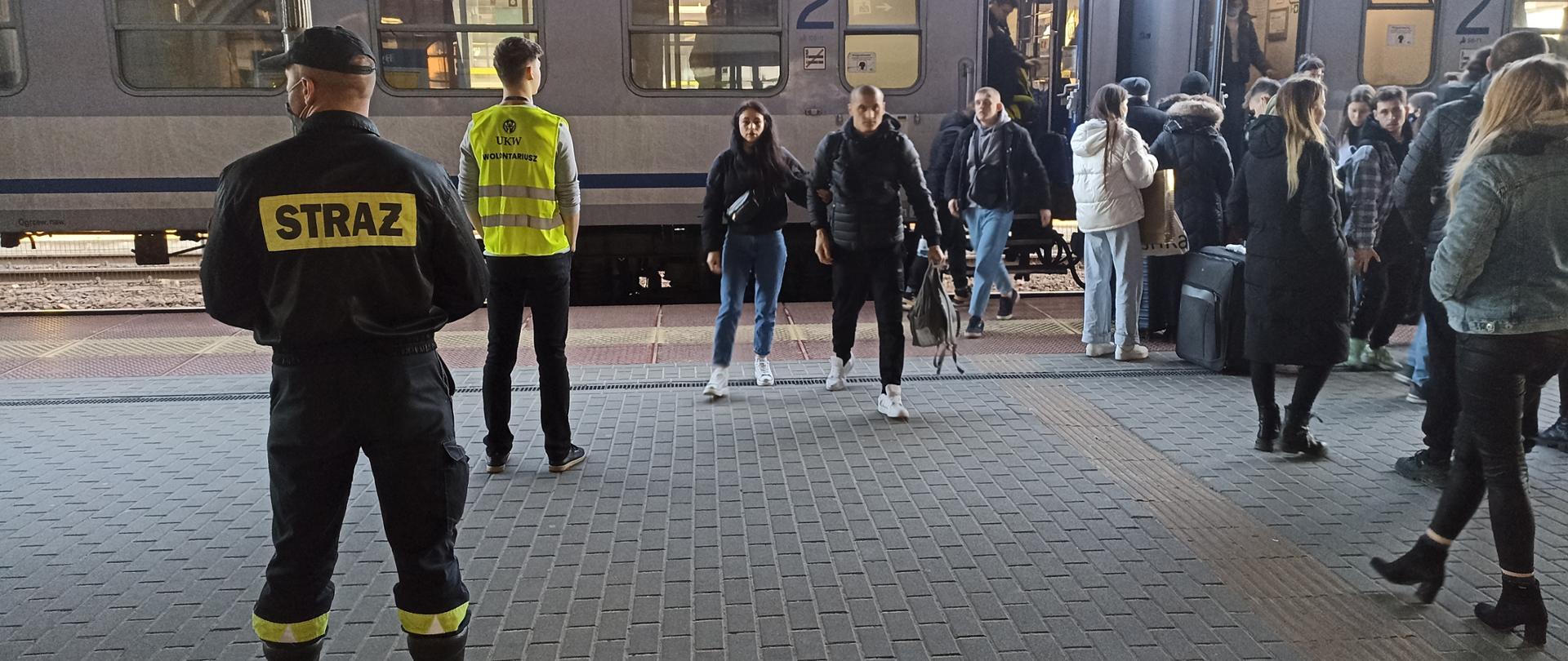 Z lewej strażak PSP w czarnym ubraniu koszarowym, Strażak stoi tyłem do osoby robiącej zdjęcie, a przodem do pociągu stojącego na peronie, z pociągu wysiada kilka osób, głównie kobiety. 