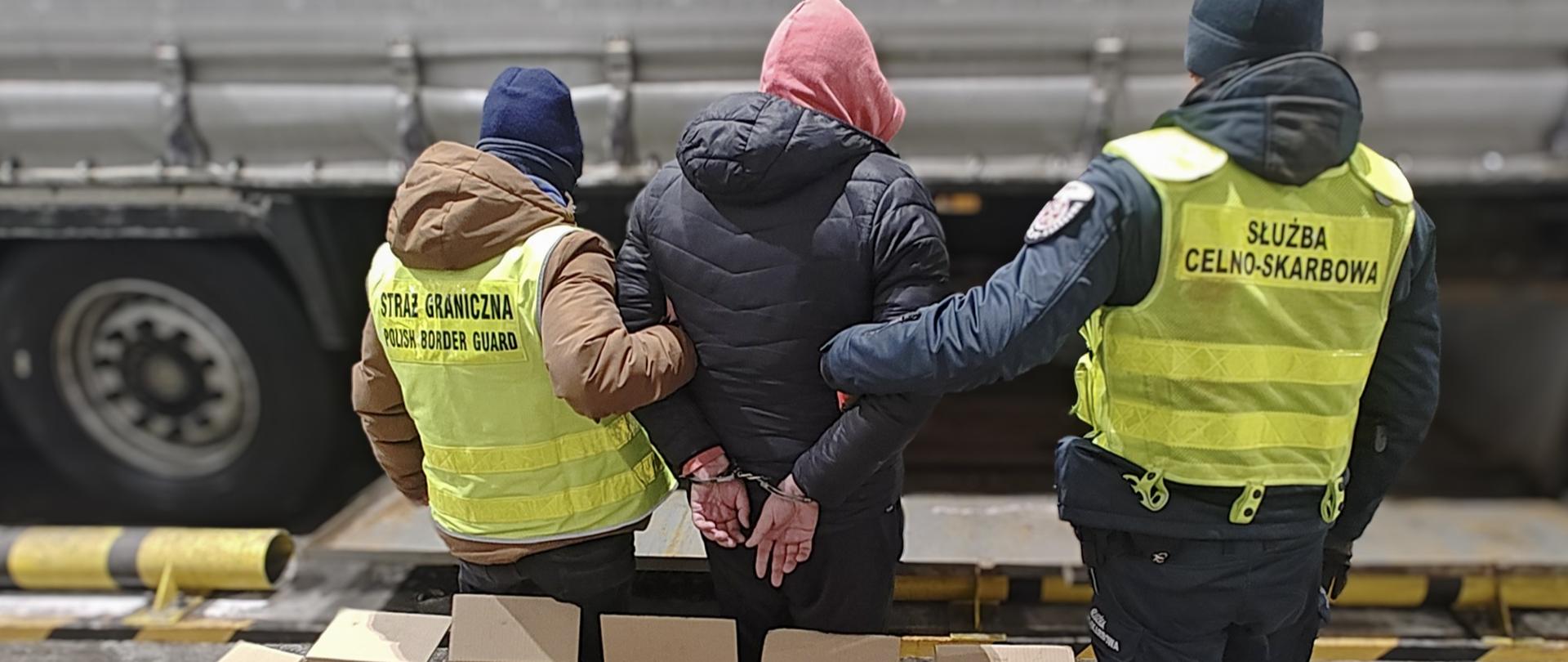 Dwie umundurowane osoby stoją przy osobie z rękami trzymanymi za plecami. Przed nimi ustawione są otwarte kartony z pakunkami.