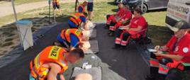 Na zdjęciu widzimy drużynę czteroosobową wykonującą reanimację na fantomach. Każdy zawodnik ma ubraną kamizelkę ratunkową element zabezpieczenia ratowników wodnych. W tle Operator Kamery TVP3 Kielce nagrywa rywalizację.
