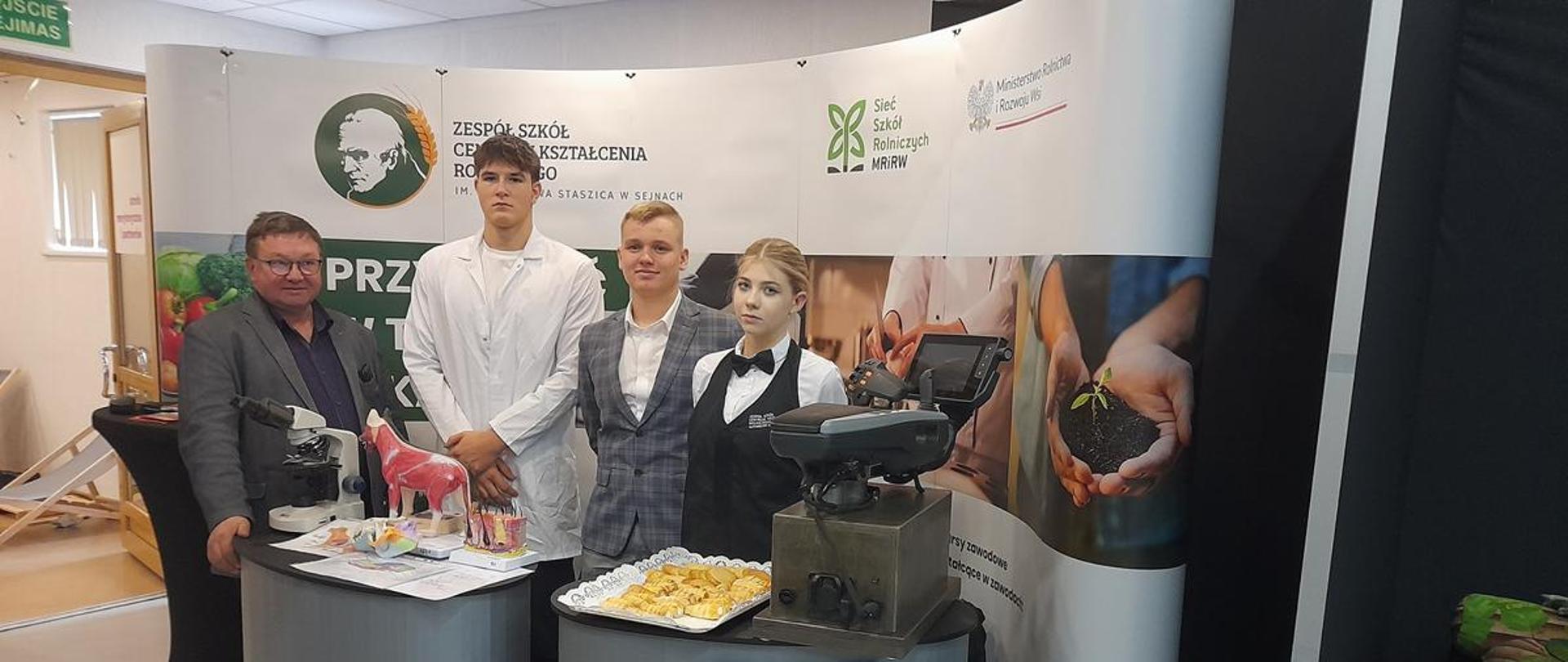 Troje uczniów i nauczyciel na stoisku promocyjnym.