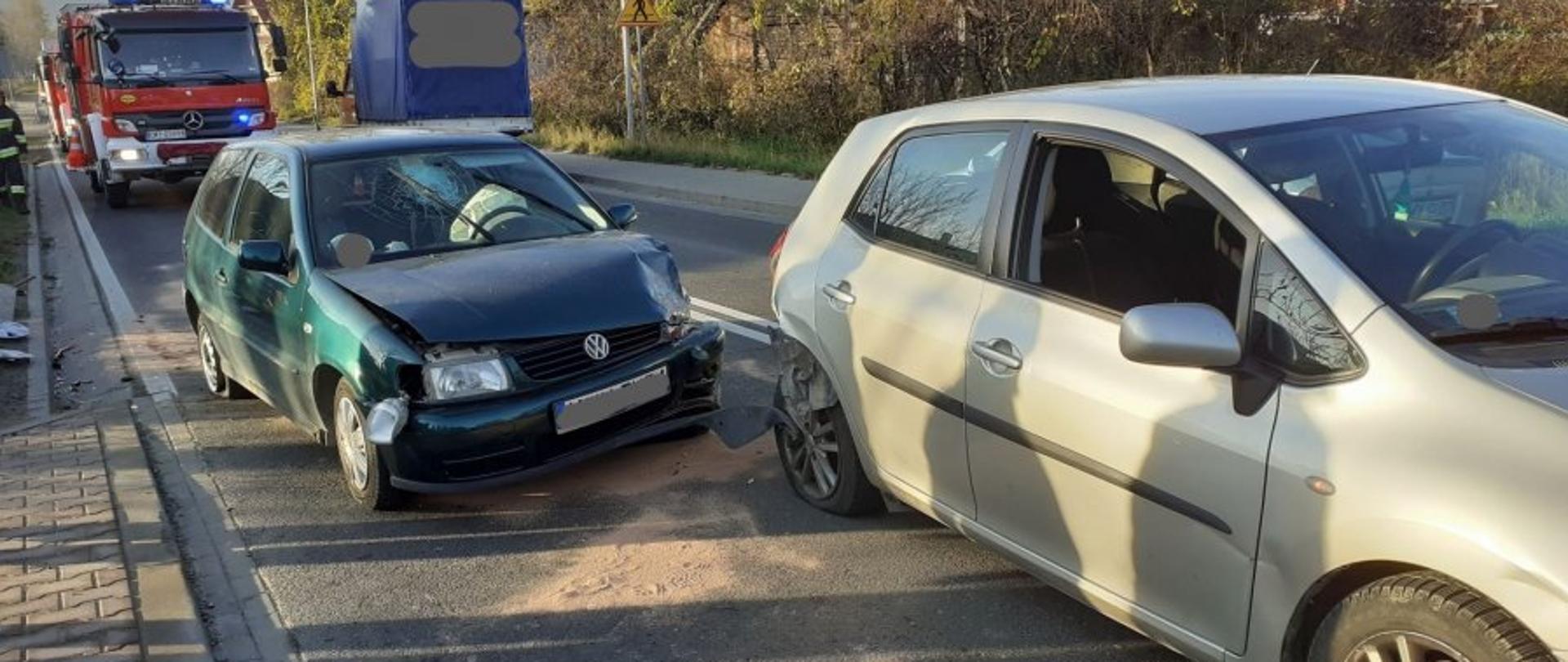 Zdjęcie przedstawia zderzenie dwóch samochodów osobowych. Na pierwszym planie z lewej strony zielony samochód z rozbitym przodem, pęknięta przednia szyba, wystrzelona poduszka powietrzna. Po prawej stronie srebrny samochód z rozbitym prawym tyłem. W dalszym planie samochód strażacki. Zdarzenie na jezdni 