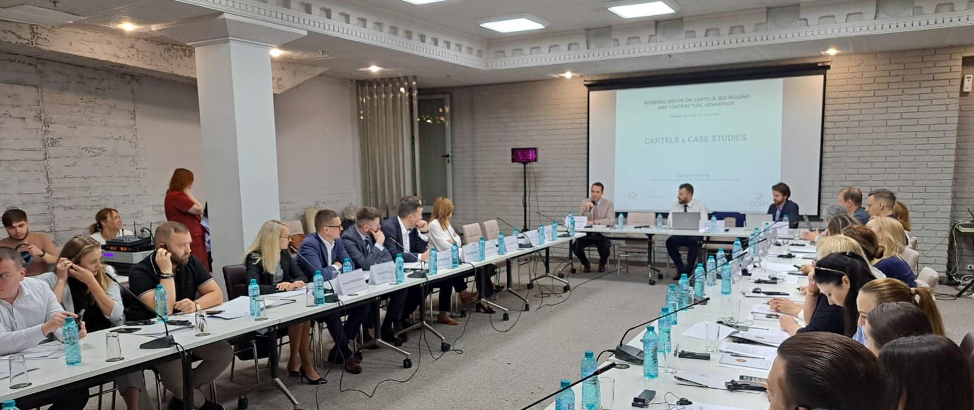 Uczestnicy warsztatów słuchają jednego z trzech prowadzących siedzących u szczytu stołu, za którym ustawiony jest ekran z wyświetlaną prezentacją