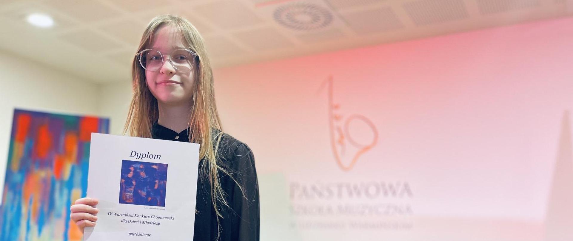 Dziewczynka stoi przy fortepianie Fazioli i trzyma w ręce dyplom.