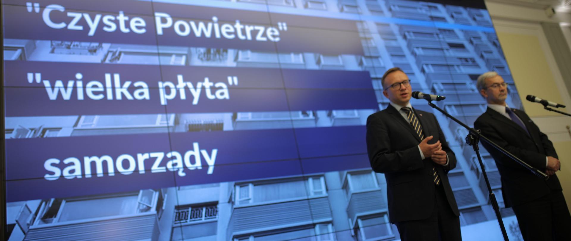 Przed mikrofonami stoją minister Jerzy Kwieciński (P) i wiceminister Artur Soboń (L) za nimi napis Czyste Powietrze, wielka płyta, samorządy