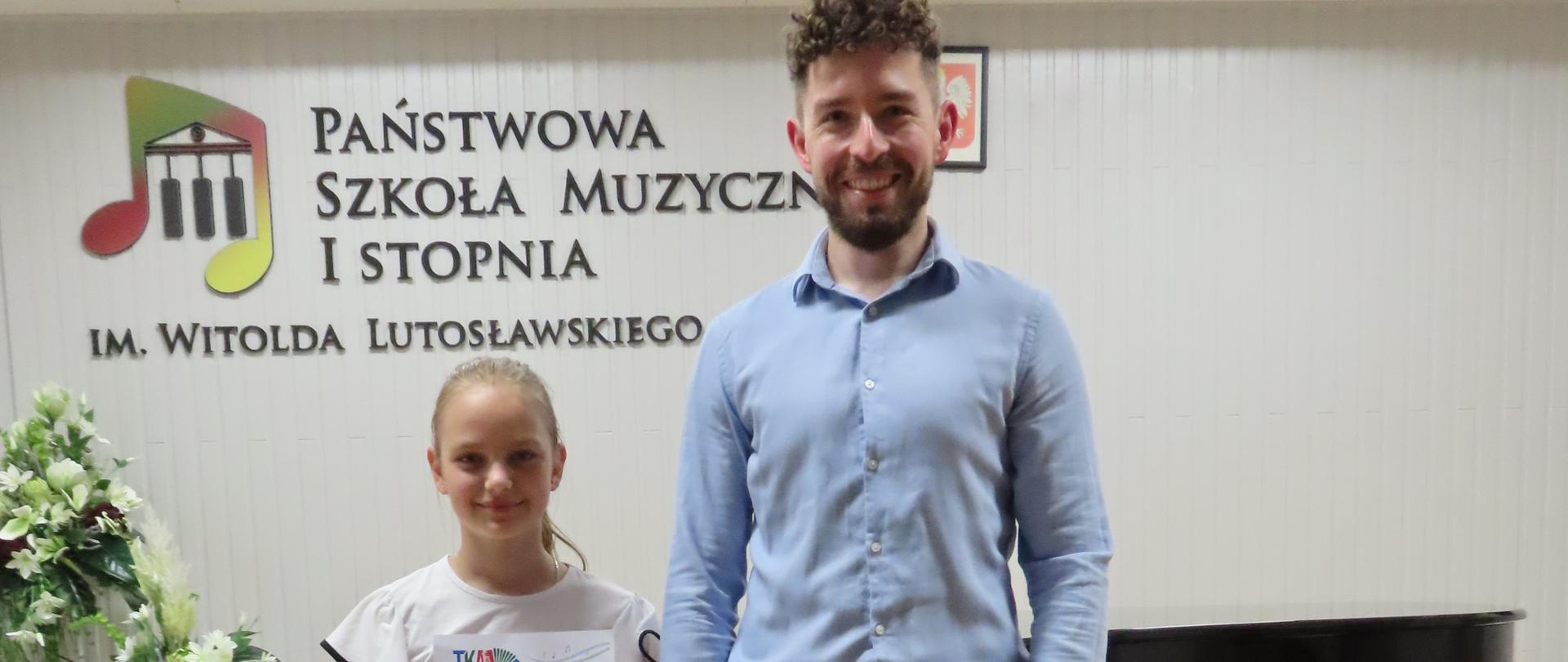 Zdjęcie przedstawia aulę państwowej szkoły Muzycznej I st. im. Witolda lutosławskiego w Jaśle. Ściany auli posiadają kolor biały, a podłoga jasnobrązowy. Na tylnej ścianie znajduje się logo szkoły oraz godło Polski. Na głównym planie widać uczennice klasy akordeonu , Milenę Żabińską wraz z nauczycielem Jakubem Zastawnym, za nimi znajduje się czarny fortepian. Uczennica trzyma dyplom wyróżnienia z konkursu akordeonowego. Na podłodze, przed uczennicą i jej nauczycielem, stoi czerwony akordeon klawiszowy.