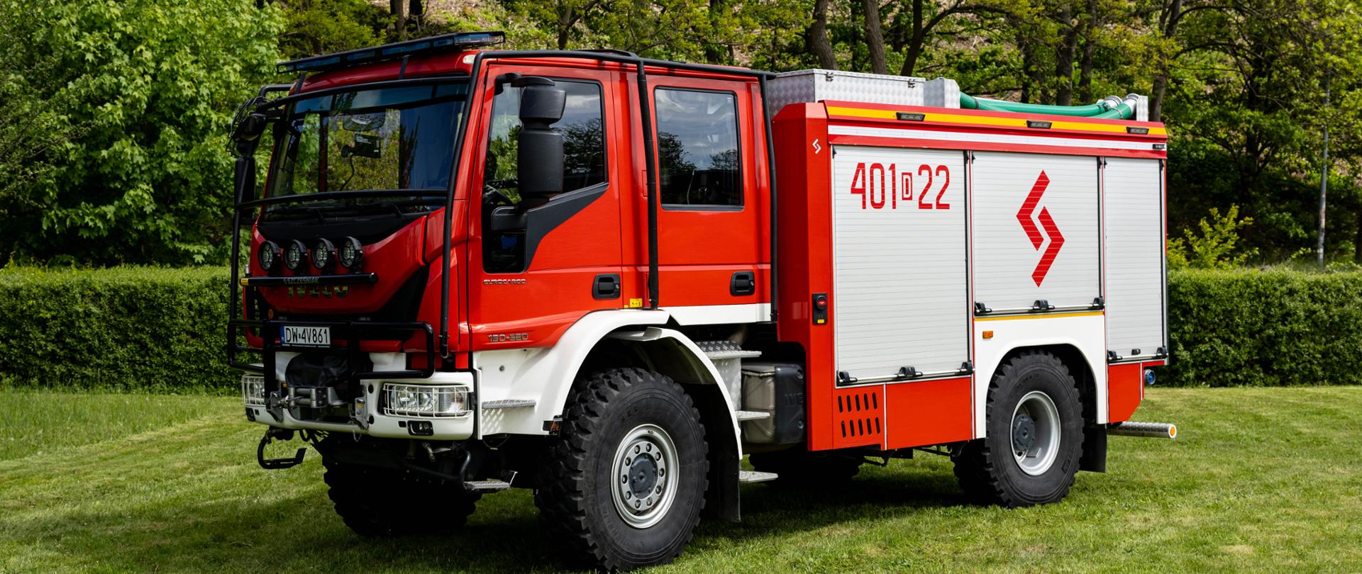 Czerwono-biały, uterenowiony samochód ratowniczo-gaśniczy marki IVECO Eurocargo na grubych, terenowych oponach. Na boku kabiny widnieje biały napis operacyjny „401[D]22”, a na żaluzji skrytki sprzętowej – czerwone, geometryczne logo. Pojazd stoi na trawiastym placu na tle zielonych drzew.