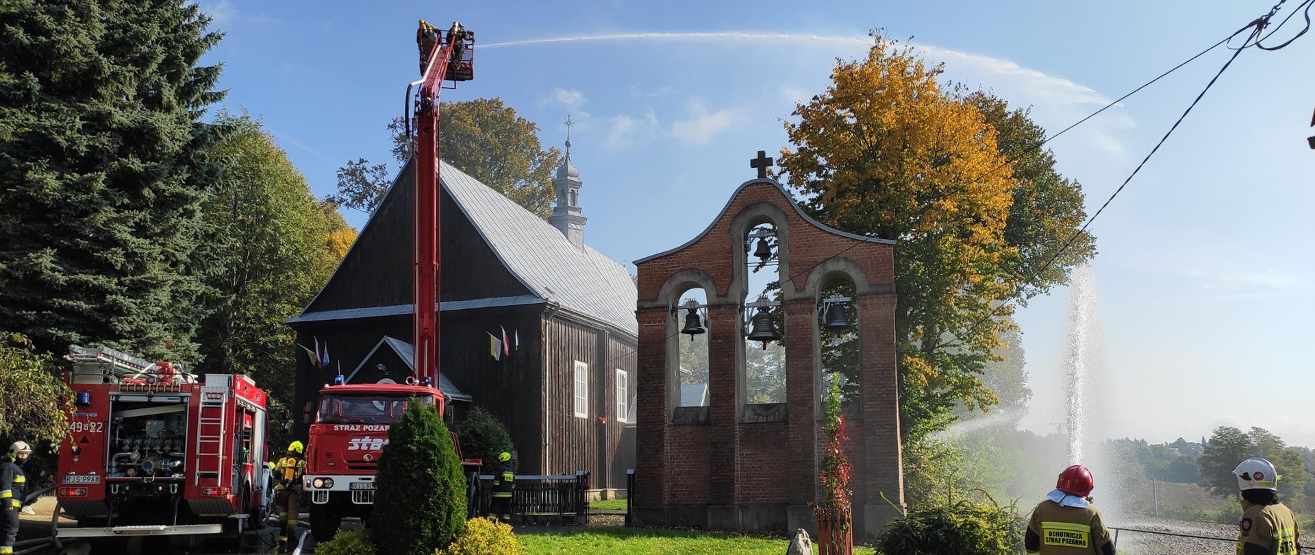 na zdjęciu zabytkowy drewniany kościół, samochód gaśniczy, podnośnik, w podnośniku strażacy podają prąd wody