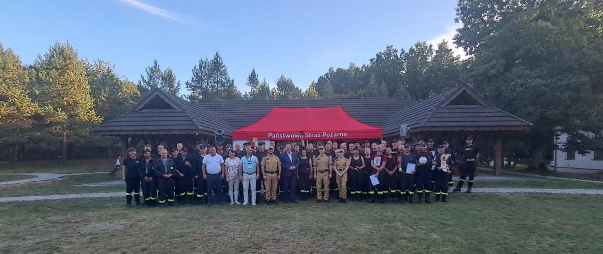 Przed czerwonym namiotem stoi kilkudziesięciu strażaków w mundurach oraz osoby w cywilnym ubraniu.