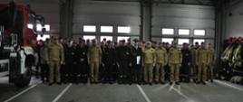 Na zdjęciu w garażu JRG Nr 1 w KP PSP w Ostrowcu Św. widać strażaków pozujących do zdjęcia grupowego po uroczystej zmianie służby. W środku grupy stoi trzech odchodzących na emeryturę strażaków. W tle samochód pożarniczy oraz ubrania strażackie 