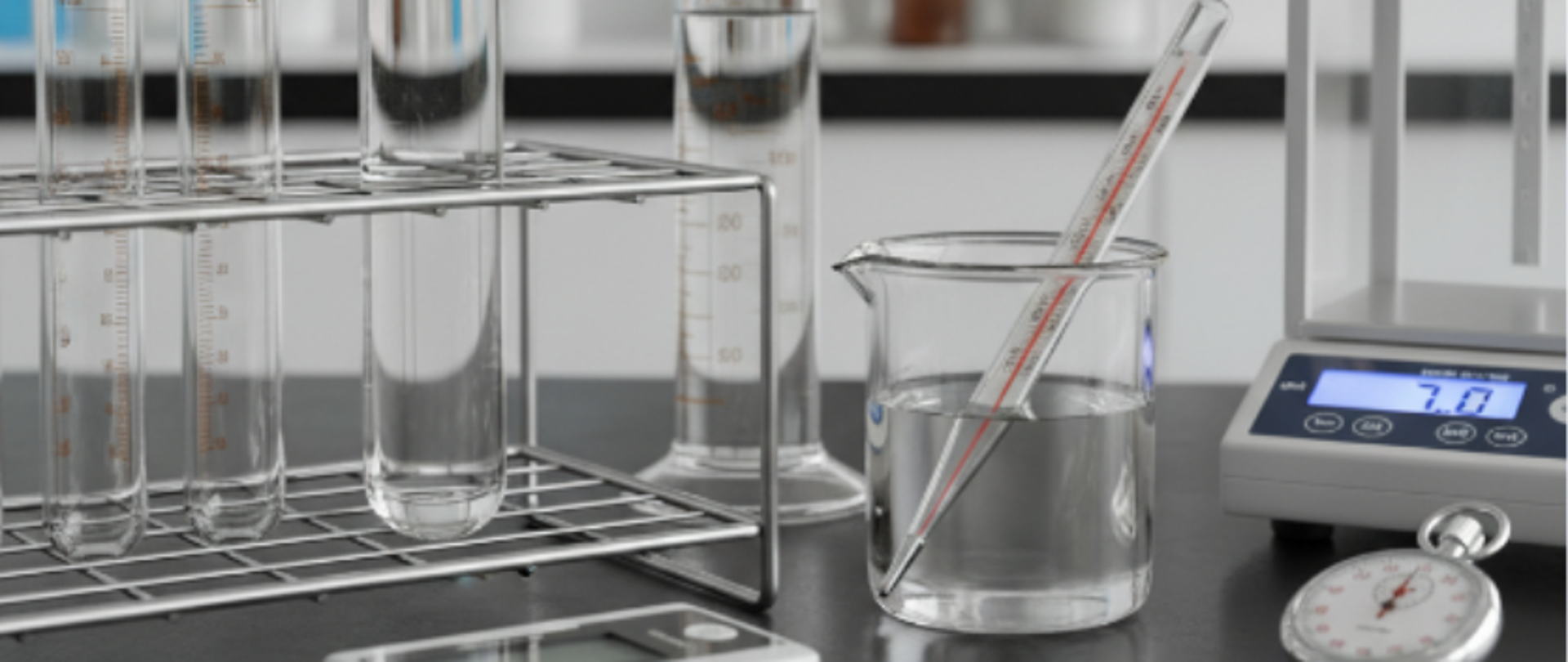 Zdjęcie przedstawia fragment laboratorium, na którym widać probówki z wodą, pojemniki z chemikaliami, termometr, wagę i czasomierz.