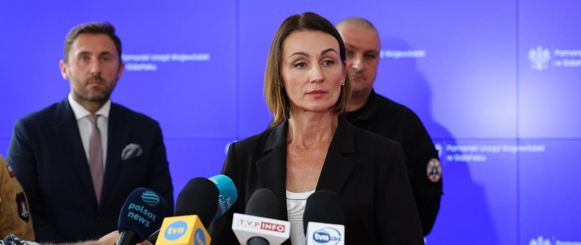 Kobieta (wojewoda) stoi przed mikrofonami i odpowiada na pytania dziennikarzy