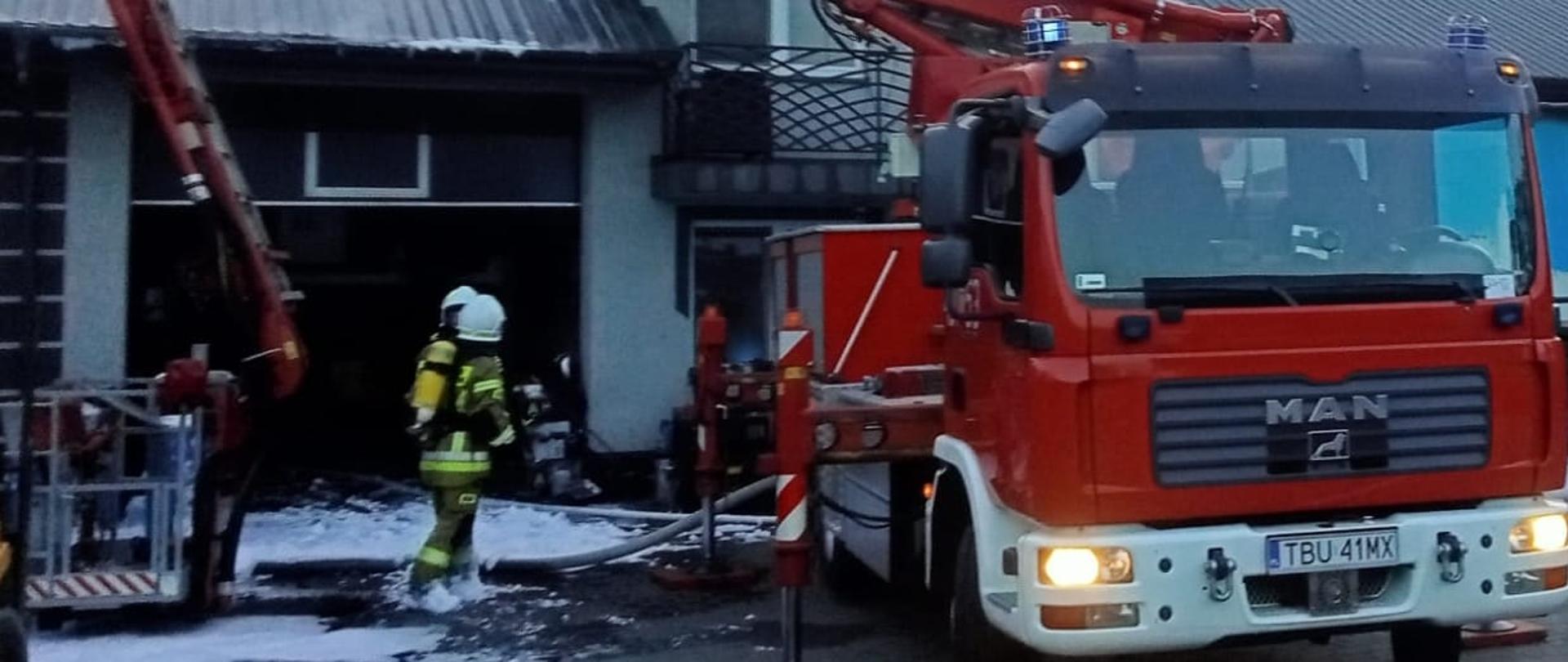 Rozstawiony podnośnik mechaniczny, stojący przed budyniem hali, w której był pożar. Obok podnośnika stoi 2 strażaków OSP zabezpieczonych w ubrania specjalne, hełmy oraz aparaty ochrony dróg oddechowych. Z dachu budynku widać wydobywający się dym. 