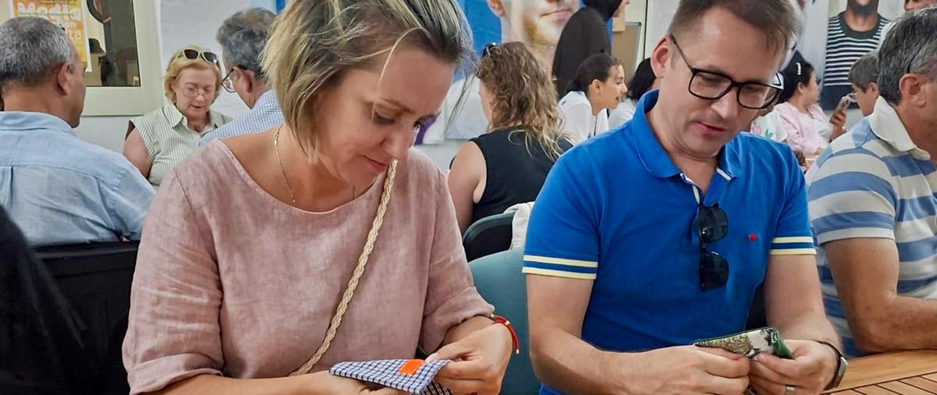 On the left, a woman is sitting at a table sewing, with a man seated to her right. Both are sewing letters. In the background, a group of people is collaborating as part of the event “The Courage to Learn.”
