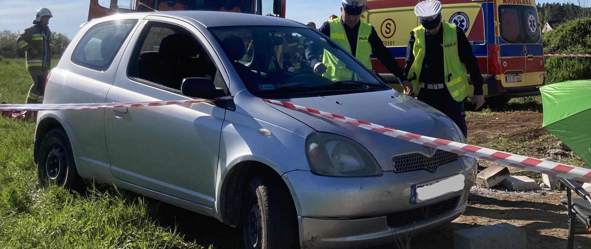 Zdjęcie przedstawia samochód osobowy, pod którym znajdowała się poszkodowana kobieta. Za nim widać fragmenty ambulansu oraz samochodu pożarniczego. Przy aucie osobowym stoją funkcjonariusze Policji.