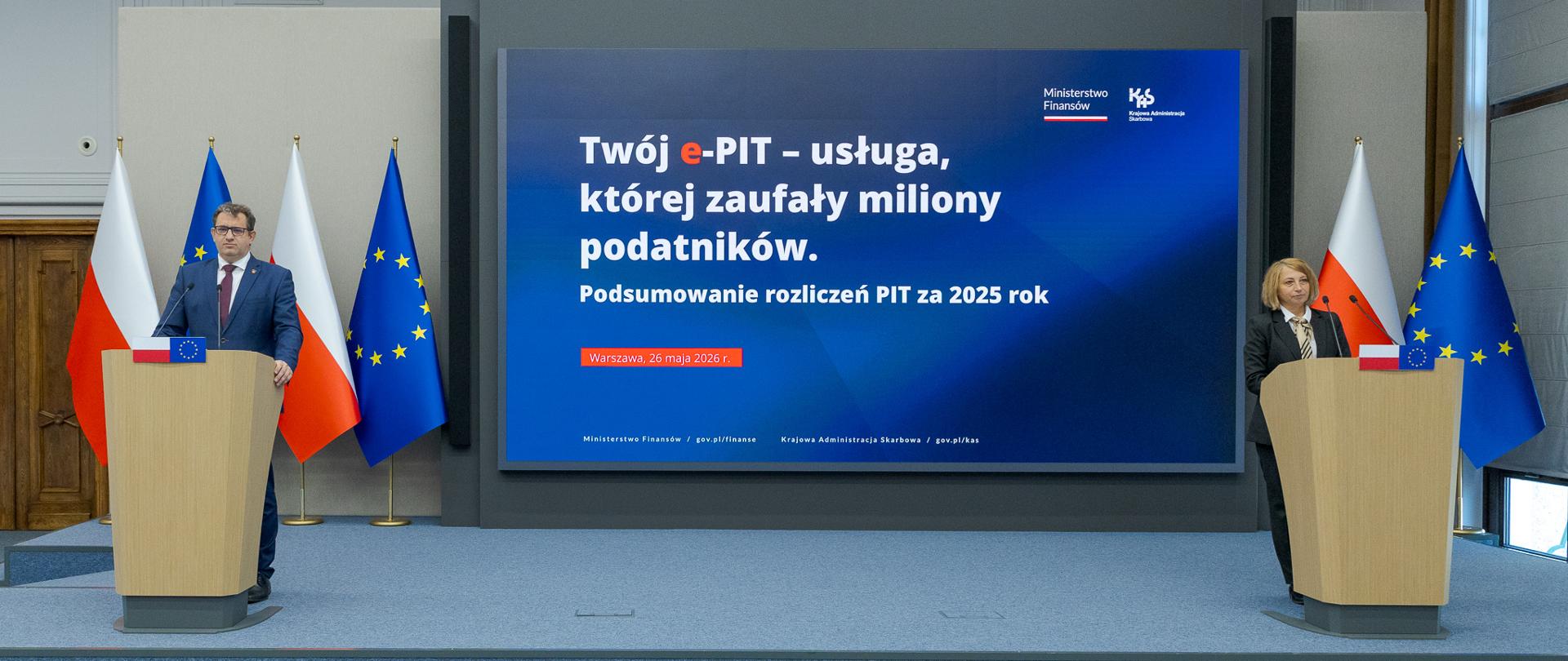Dwie osoby stoją przy oddzielnych mównicach, a między nimi ekran z napisem „Twój e‑PIT – usługa, której zaufały miliony podatników” oraz informacją o podsumowaniu PIT za 2025 rok.
