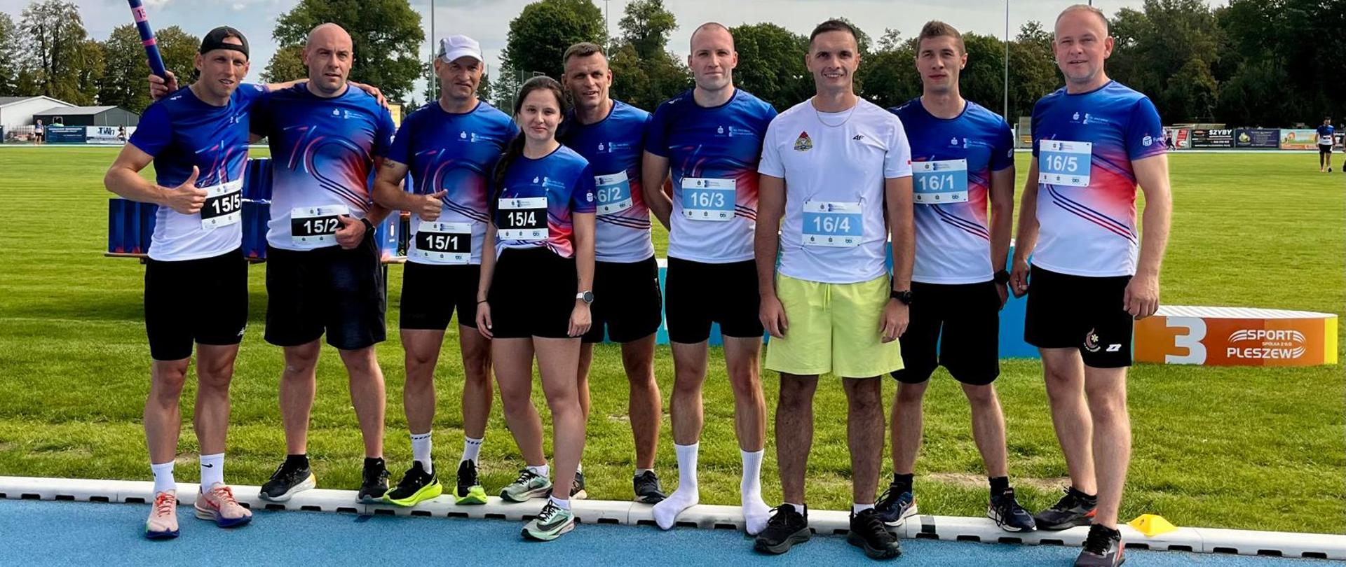 Grupa ośmiu biegaczy (jedna kobieta i siedmiu mężczyzn) pozuje razem do zdjęcia na niebieskiej bieżni lekkoatletycznej. Większość z nich ma na sobie niebiesko-białe koszulki sportowe z numerami startowymi oraz czarne spodenki. W tle widać murawę boiska i podium.