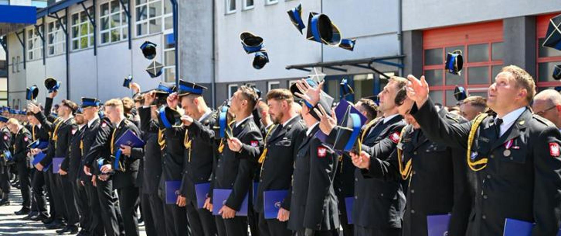 Grupa strażaków Państwowej Straży Pożarnej w galowych mundurach stoi w szeregu na placu przed budynkiem komendy. Wszyscy z uśmiechem i radością podrzucają w górę swoje czapki rogatywki podczas uroczystego apelu. W rękach trzymają niebieskie foldery lub dyplomy. W prawym dolnym rogu widnieje podpis: „st. str. kdt Piotr Krzak | Państwowa Straż Pożarna”.