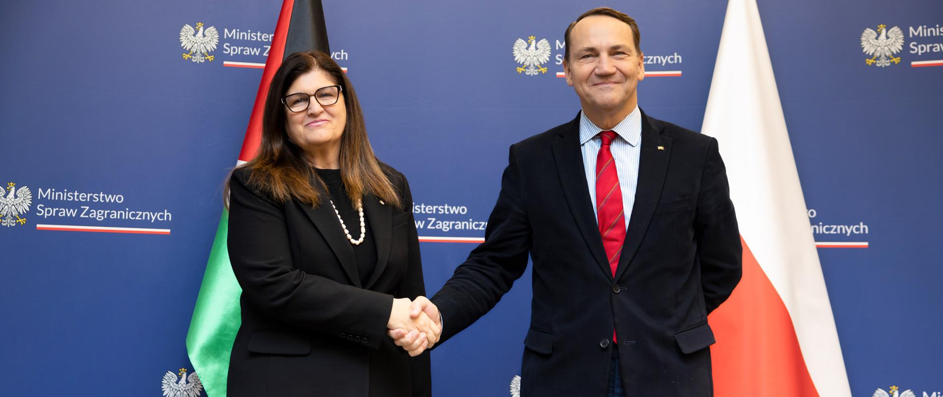 Ministrowie Spraw Zagranicznych Polski i Palestyny stoją przed ścianką z logo Ministerstwa Spraw Zagranicznych, na tle flag obu państw, i podają sobie ręce. Oboje są ubrani w formalne stroje i patrzą w stronę aparatu, pozując do oficjalnego zdjęcia.
