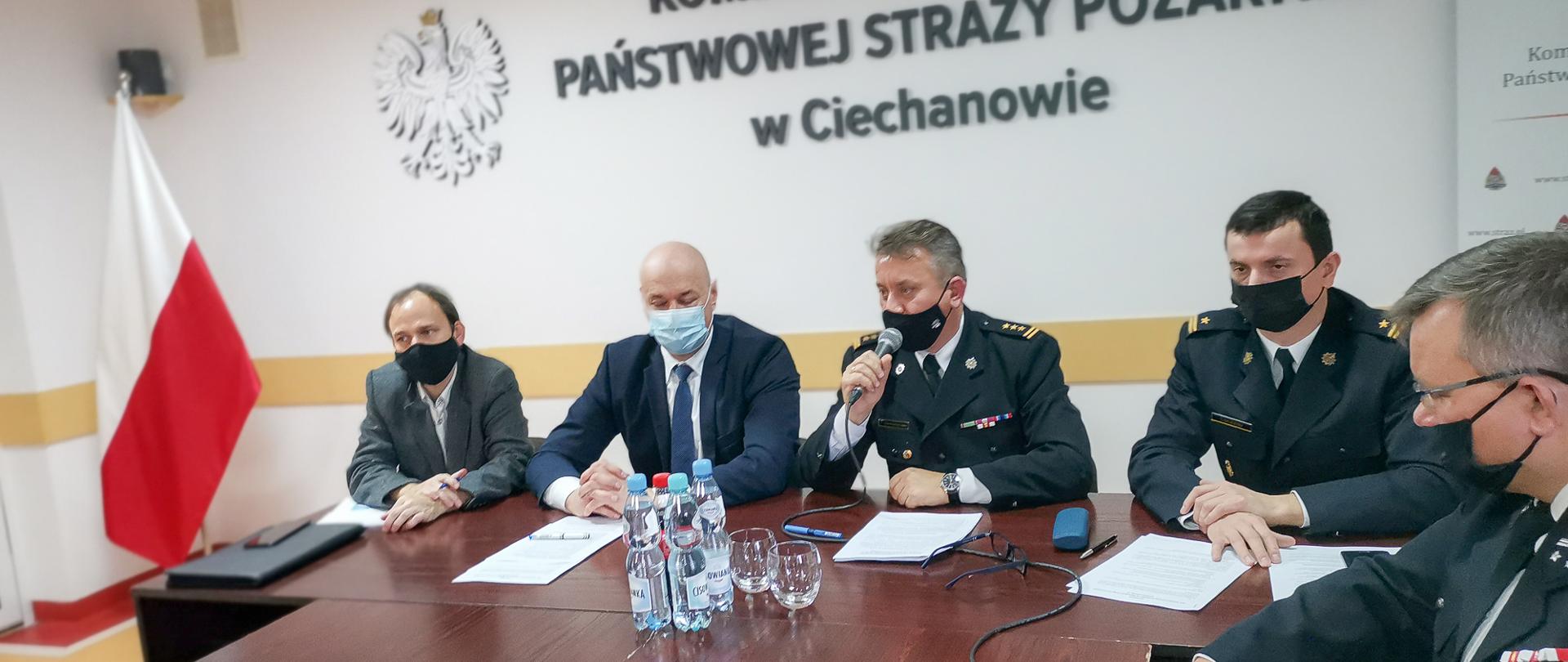 Zdjęcie przedstawia pięć osób na spotkaniu roboczym dotyczącym szczepień przeciwko COVID-19. Strażak w środku trzyma mikrofon i przemawia. W tle godło RP na ścianie. Po lewej stronie zdjęcia stoi flaga Polski.
