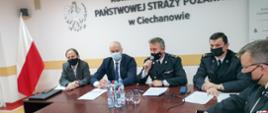 Zdjęcie przedstawia pięć osób na spotkaniu roboczym dotyczącym szczepień przeciwko COVID-19. Strażak w środku trzyma mikrofon i przemawia. W tle godło RP na ścianie. Po lewej stronie zdjęcia stoi flaga Polski.