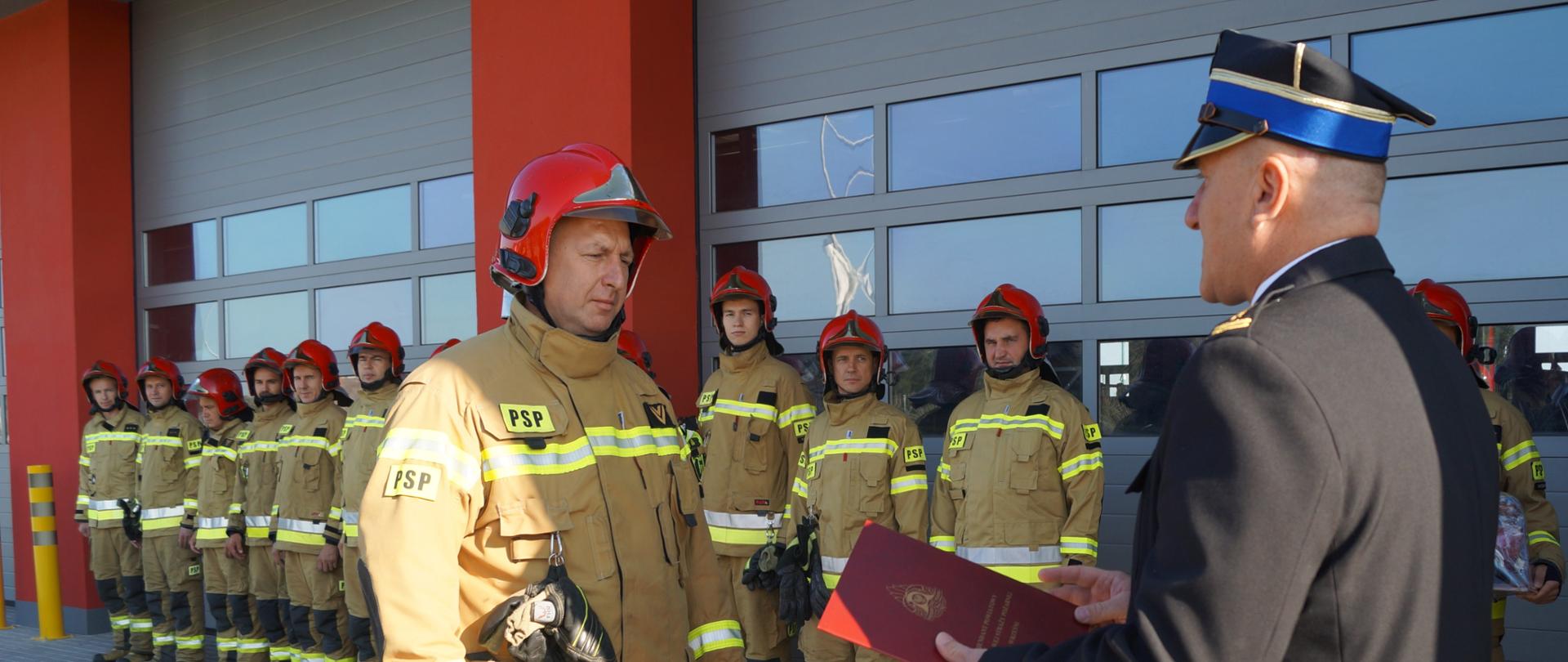 Zdjęcie przedstawia strażaka i Komendanta Powiatowego. W tle pozostali strażacy