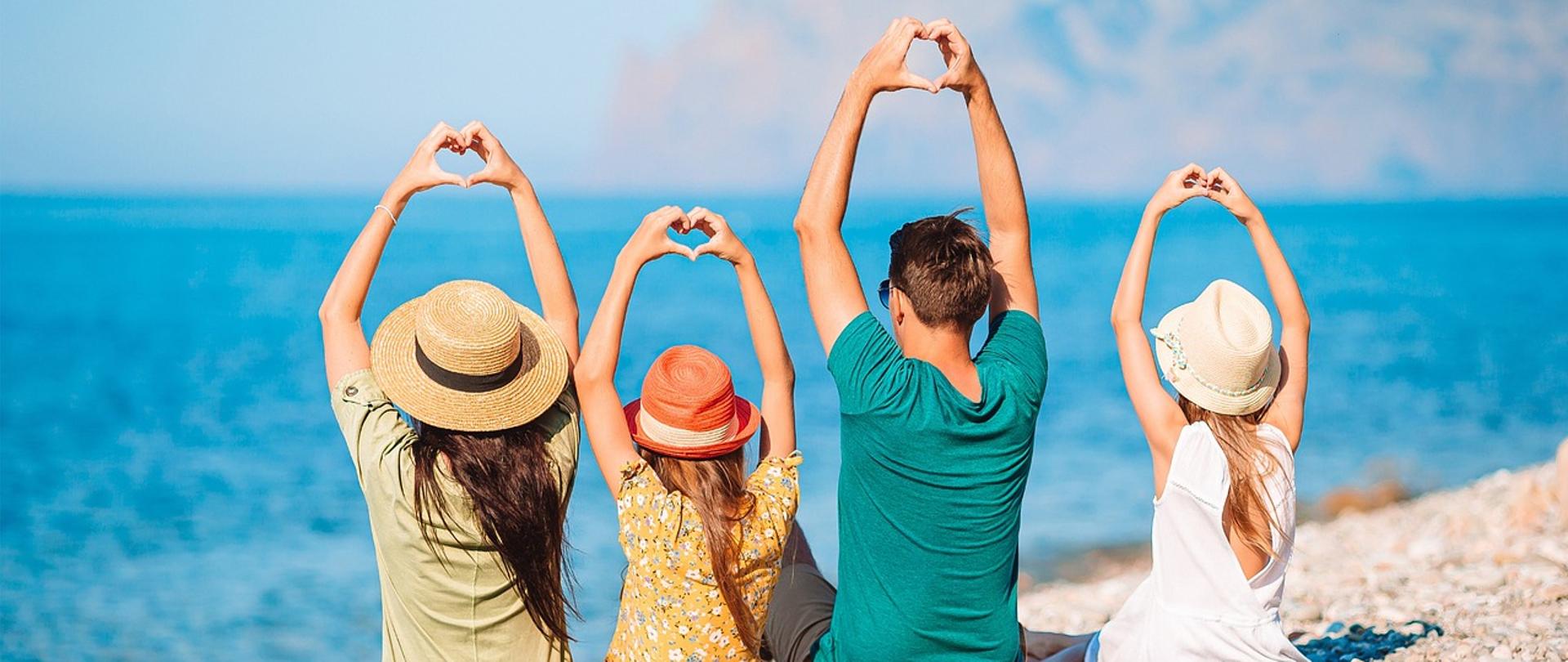 Rodzina siedząca na kamienistej plaży tyłem do obiektywu – dwie kobiety, mężczyzna i dziewczynka – wszyscy w letnich ubraniach i słomkowych kapeluszach. Trzymają ręce uniesione nad głową, tworząc kształt serca palcami. W tle błękitne morze i zamglone górskie wybrzeże. Letni, pogodny dzień.