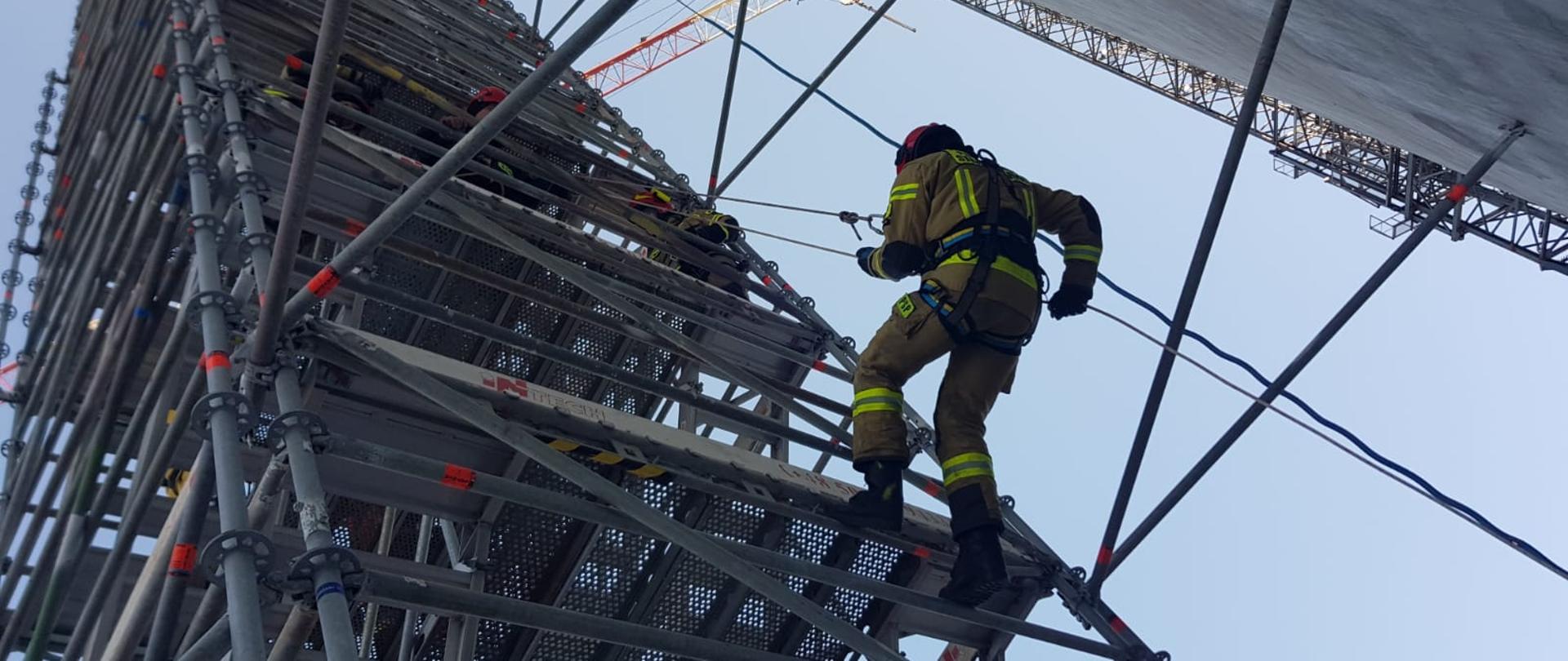 Strażak w mundurze bojowym i szelkach bezpieczeństwa korzystając z przyrządu zjazdowego zjeżdża po linie po zewnętrznej stronie klatki schodowej w przestrzeni pomiędzy klatką a ścianą zbiornika. 