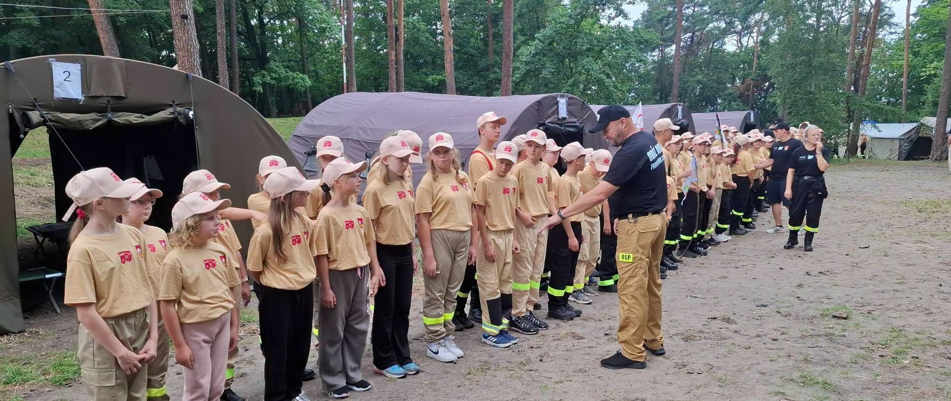 W dwuszeregu ustawiona grupa dzieci i młodzieży na obozie. W tle duże namioty i las.