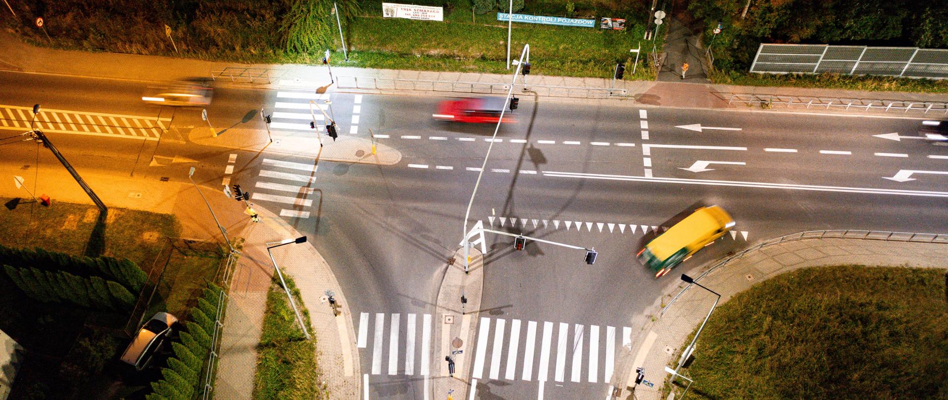 Widok z lotu ptaka na skrzyżowanie ulic nocą, z pojazdami i przejściami dla pieszych. Oświetlenie podkreśla ruch samochodów, z jednym samochodem w ruchu, co wskazuje na prędkość. Kontrast między ciemnym otoczeniem a oświetloną drogą tworzy dynamiczny efekt wizualny. Obraz uchwyca esencję życia miejskiego po zmroku, podkreślając ruch i infrastrukturę miejską.