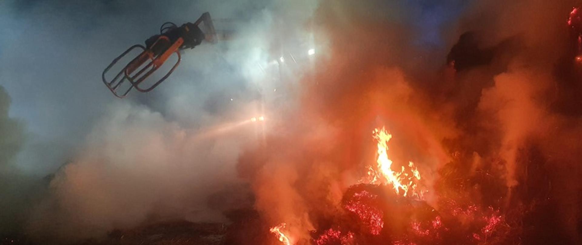 Zdjęcie przedstawia pożar balotów w Nowych Kiejkutach, z za ognia widać ciągnik rozciągający baloty w celu przelania wodą.