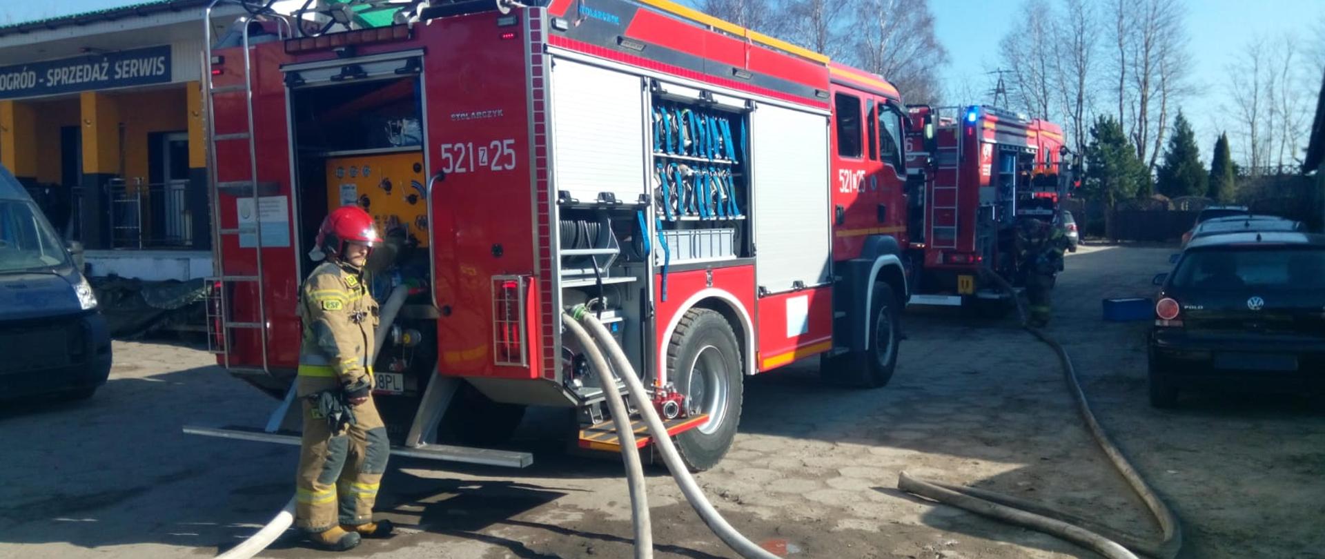 Dwa wozy straży pożarnej ustawione wzdłuż drogi, strażak obsługuje linię gaśniczą przy tylnej części pojazdu podczas prowadzonej akcji.