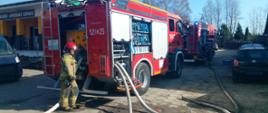 Dwa wozy straży pożarnej ustawione wzdłuż drogi, strażak obsługuje linię gaśniczą przy tylnej części pojazdu podczas prowadzonej akcji.