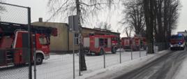 Na zdjęciu widać budynek Szkoły Podstawowej w Sulisławicach. Budynek jest w kolorze żółto-zielonym. Przed budynkiem stoją samochody straży pożarnej. Od lewej są to: drabina mechaniczna, dwa ciężkie wozy gaśnicze oraz jeden średni wóz gaśniczy. Pojazdy posiadają sygnalizację ostrzegawczą i są w kolorze czerwonym. Przed nimi widoczne jest ogrodzenie szkoły wzdłuż, którego są posadzone drzewa. Zdjęcie zostało wykonane w porze zimowej, w ciągu dnia. Na pierwszym planie widać jezdnię, przy której znajduje się znak drogowy.