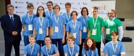 Na tle ściany z logotypami i banerem, grupa kilkunastu młodych ludzi i dwóch dorosłych mężczyzn pozuje do grupowego zdjęcia. Większość młodych osób ubrana jest w jasnoniebieskie koszulki z różnymi nadrukami i akredytacjami na szyjach. Niektórzy stoją, a niektórzy klęczą w przednim rzędzie.
Po lewej stronie, na stojąco, znajduje się starszy mężczyzna w ciemnym garniturze i okularach. Po prawej stronie, również na stojąco, jest mężczyzna z brodą i okularach, ubrany w zieloną koszulkę i białą koszulę w kratę.
Za grupą widać baner z napisem „BOI” i tekstem „Baltic Olympiad in Informatics”. Ściana za nimi zawiera powtarzające się logotypy różnych organizacji. Oświetlenie jest jasne, prawdopodobnie wewnątrz budynku. Atmosfera sugeruje wydarzenie edukacyjne lub konkursowe, takie jak olimpiada informatyczna.